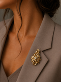 A close-up of a woman wearing a textured gold floral brooch with a small clear center stone, pinned to her taupe blazer.