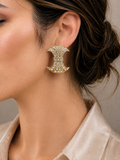 A close-up of a woman's ear wearing a gold crescent-shaped earring adorned with clear rhinestones, set against a neutral beige background.