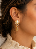 A close-up of a woman's ear wearing a gold drop earring with a large white pearl and small clear crystals.