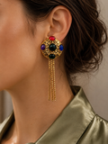 A close-up of a model wearing a gold dangle earring with colorful gemstone accents and hanging chains, paired with a satin olive green shirt.