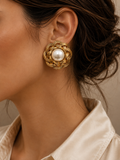 A close-up of a woman wearing a gold-toned woven flower-shaped earring with a white pearl at the center, paired with a cream-colored collared shirt.