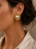 A close-up of a woman wearing a gold-toned rope-shaped earring with a white pearl center, paired with a cream satin shirt.