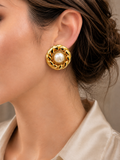 A close-up of a woman wearing a round gold earring with a central white pearl and textured gold detailing.