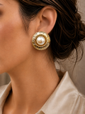 A close-up of a model wearing a round gold earring with a textured halo and a central white pearl.