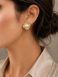 A close-up of a model's ear wearing a gold-toned square stud earring with a central white pearl surrounded by small clear stones.