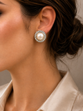 A close-up side profile of a woman wearing a round stud earring with a white pearl surrounded by a halo of small clear crystals, paired with a light beige satin collared shirt.