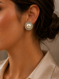 A close-up of a woman's ear wearing a round stud earring with a white pearl surrounded by a halo of small clear stones.