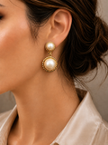 A close-up of a woman's ear wearing a gold and pearl drop earring, with her hair pulled back and a light-colored satin shirt visible.
