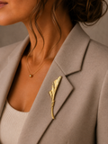 A close-up of a woman wearing a gold leaf-shaped brooch pinned to her taupe blazer, paired with a delicate gold necklace.
