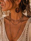 A close-up lifestyle shot of a woman wearing bohemian jewelry, featuring a large circular face pendant earring and layered beaded necklaces in earthy tones.